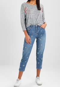 Maglietta grigia a maniche lunghe con ricami floreali; jeans blu chiaro con decorazioni floreali bianche; sneakers bianche; fit comodo, stile casual.