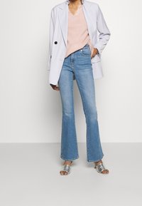 Hellgrauer doppelreihiger Blazer über einem rosa V-Ausschnitt-Pullover, kombiniert mit hellblauen ausgestellten Jeans und silbernen Sandalen mit Absatz.