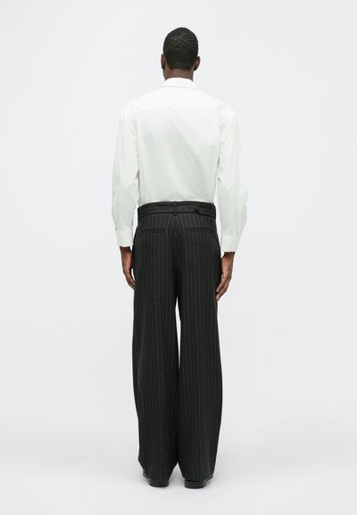 Witte blouse met lange mouwen, gecombineerd met een ruimvallende, donker gestreepte pantalon. De stof lijkt soepel met duidelijke naden en zakken.