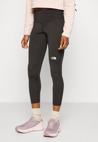 Leggings de sport noirs avec une taille haute, arborant un logo blanc. Associés à des chaussures de sport violettes claires et des chaussettes de couleur crème.