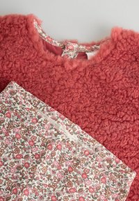 Czerwony sweter o fakturowanej powierzchni z puszystym materiałem, zestawiony z tkaniną w kwiatowy wzór z różowymi i brązowymi kwiatami na jasnym tle.