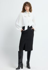 Blouse blanche avec un nœud, manches volumineuses, associée à une jupe crayon noire avec une fente avant, et des bottes noires en cuir crocodile jusqu'aux genoux.