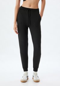Schwarze Jogginghosen mit Kordelzugbund, Seitentaschen, elastischen Bündchen und glatter Textur; mit Paneel-Details an den Beinen.