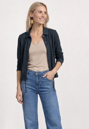 Femme blonde souriante portant un haut beige, une chemise sombre boutonnée, un jean bleu taille haute et de grandes boucles d'oreilles décoratives.