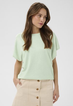 Kobieta z długimi brązowymi włosami, ubrana w jasnorozmowiony zielony top z krótkim rękawem i beżową spódnicę na guziki, z rękami w kieszeniach, patrząca w prawo.