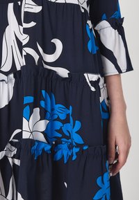 Vestido florido em camadas com cores azul-marinho, branco e azul. Feito de um tecido leve, com detalhes em babados e uma textura suave.