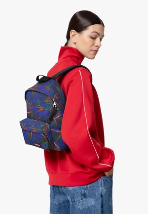 Mochila con base azul, patrón de hojas verdes y rojas, forma rectangular, correas acolchadas y bolsillo frontal con cremallera. Llevada sobre un sudadera roja.
