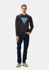 Felpa nera con un logo a triangolo blu sul fronte, abbinata a jeans blu scuro e sneakers grigie con suole in gomma.