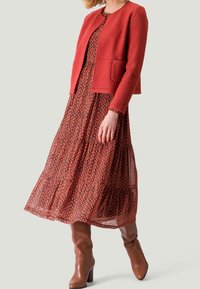 Červená texturovaná bunda s kulatým výstřihem a kapsami, vrstvená přes vzorované dlouhé splývavé šaty se zaoblenými motivy v odstínech červené a černé.