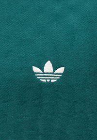Λευκό λογότυπο Adidas trefoil κεντημένο σε πετρόλ πλεκτό ύφασμα, κεντραρισμένο και καθαρά ορατό.