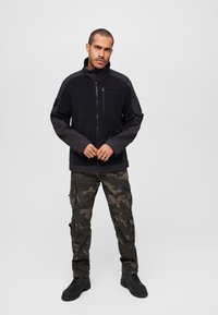 Black fleece jacket with a zippered front and high collar, paired with camo cargo pants featuring multiple pockets, worn with black boots.