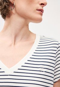 Μπλουζάκι με V λαιμόκοψη σε λευκό και ναυτικό μπλε, φτιαγμένο από μαλακό ύφασμα. Σημαντική κεντημένη λεπτομέρεια στην αριστερή πλευρά του στήθους.