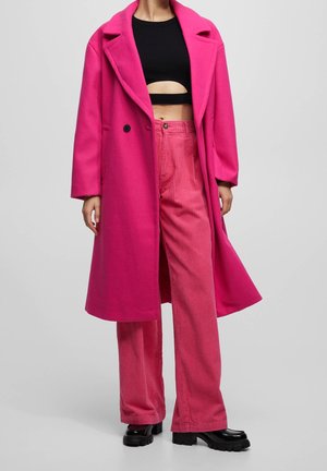 Femme portant un manteau long rose vif sur un pantalon en velours côtelé rose et un crop top noir avec des chaussures noires épaisses sur un fond uni.