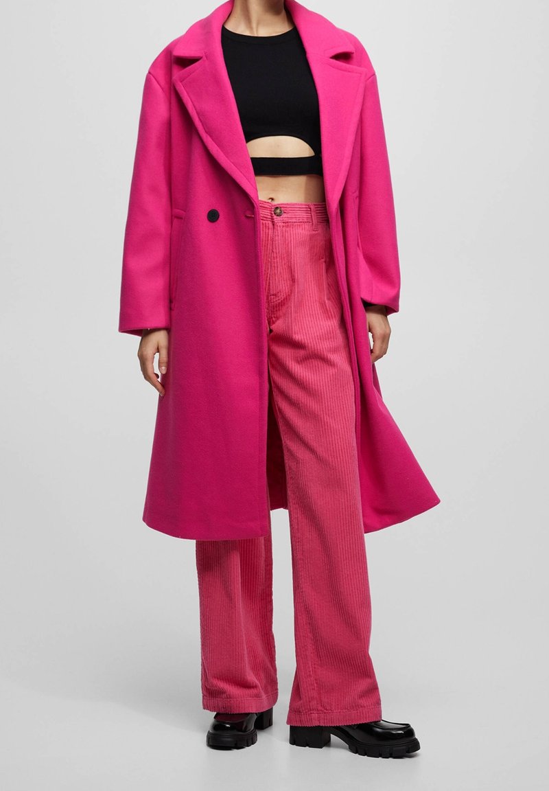 Femme portant un manteau long rose vif sur un pantalon en velours côtelé rose et un crop top noir avec des chaussures noires épaisses sur un fond uni.