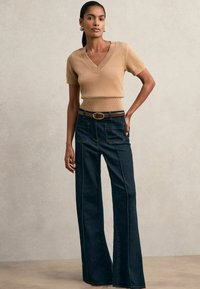 Beige, rövid ujjú, V nyakú kötött felső, sötétkék, magas derekú, bő szárú farmernadrággal párosítva. A farmernadrág kontrasztos varrással és elülső zsebekkel rendelkezik.
