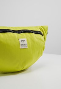Sac banane Anello vert vif avec fermeture éclair noire et petite étiquette de marque blanche, posé sur une surface blanche sur fond gris uni.