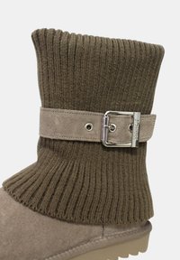 Stivaletto marrone in maglia con bordo a costine, caratterizzato da una cinghia in suede beige e dettaglio con fibbia argentata. La suola è strutturata e resistente, adatta a diverse superfici.