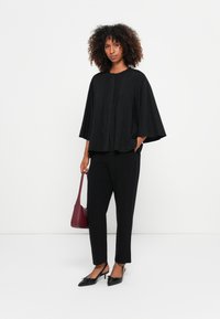 Top preto de mangas morcego com um tecido texturizado, apresentando um decote redondo e fecho na frente, combinado com calças pretas justas e sapatos de bico fino.