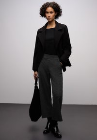 Manteau noir sur mesure, haut noir ajusté et pantalon gris texturé à jambes larges. Bottes noires et sac noir avec un détail circulaire.
