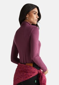 Langärmliges lila Oberteil mit hohem Kragen, getragen mit einem gemusterten rosa Schal, der tailliert über dunklen Hosen gebunden ist. Glatte Textur, figurbetontes Design.