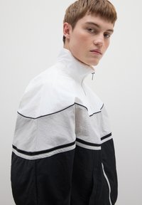 Herenjas met rits en kleurblokken in wit, grijs en zwart. Lichtgewicht stof met horizontale strepen en ritszakken.