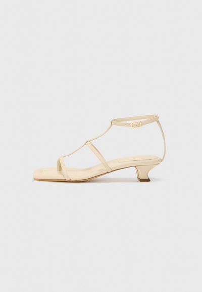 Beige heeled sandals with a T-strap design, square toe, adjustable ankle strap, and a low, block heel. Smooth leather material.