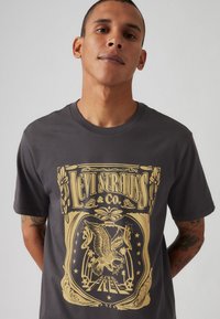 Tricou din bumbac gri, cu un design grafic mare, complicat, de culoare aurie, reprezentând o vultur și elemente decorative, cu decolteu la baza gâtului.