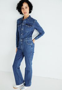 Combinaison en denim bleu foncé avec un corsage ajusté, des manches longues et deux poches poitrine. Présente des coutures contrastantes et des jambes larges.