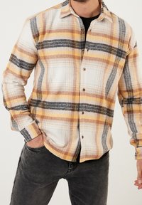 Camisa de cuadros con colores blanco, amarillo, marrón y negro. Confeccionada en tejido suave, con cierre de botones y mangas largas. Diseño casual.