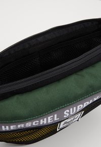 Groene en zwarte Herschel Supply pouch met rits toont de open binnenkant met zwarte gaasvakjes en stoffen voering.