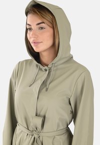 Impermeable beige avec capuche, cordon de serrage, boutons à l'avant, ceinture nouée, et une texture lisse et résistante à l'eau. Design minimaliste sans motifs.