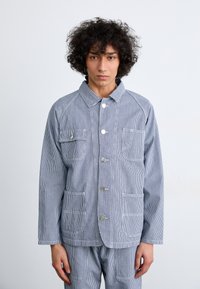 Blue and white striped cotton jacket featuring a button closure, two breast pockets, and contrasting white stitching. Simple, classic design.