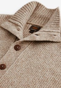 Beige Wollpullover mit geripptem Kragen, zwei braunen Knöpfen und einem strukturierten Muster. Mit einem Etikett im Kragen.