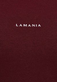 Bordowy prążkowany materiał z białym logo "LAMANIA" na środku. Faktura jest gładka z jednolitym wzorem dzianiny.