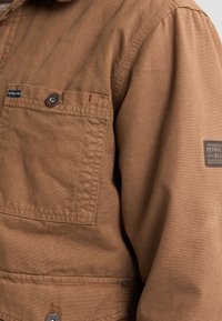 Brown canvas jacket with a chest pocket, button closures, and stitched detailing. Features a brand tag on the sleeve.
