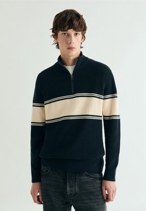Suéter azul marino con cremallera y textura acanalada, con una franja horizontal crema acentuada por dos líneas negras a lo largo del pecho.