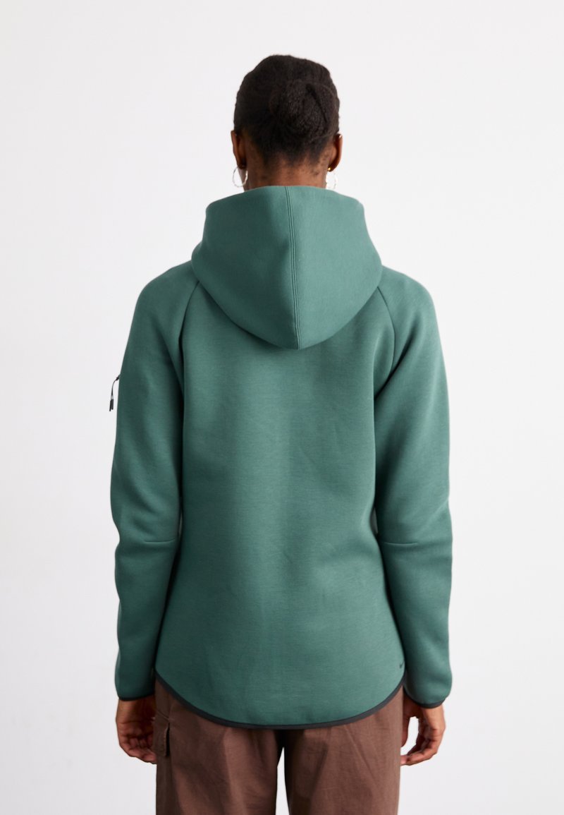 Sweat-shirt à capuche verte avec une texture lisse, une base arrondie et des poches latérales, présentant un design ajusté et un accent zippé sur la manche.