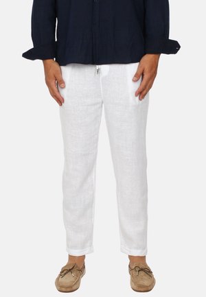 Uomo con pantaloni di lino bianchi, mocassini beige e camicia a maniche lunghe blu scuro con polsini arrotolati, in piedi contro uno sfondo semplice.