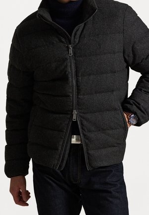 Man wearing dark gray quilted zip-up jacket over navy turtleneck and dark jeans, showing wristwatch on left wrist.
