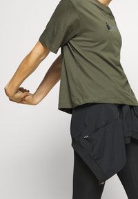 Camiseta de manga corta verde oliva con un logo negro, llevada sobre pantalones cortos negros que tienen bolsillos con cremallera y un ajuste relajado.