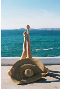 Cappello di paglia a tesa larga davanti a gambe scoperte che indossano sandali marroni decorati, con l'oceano blu sullo sfondo sotto un cielo sereno.