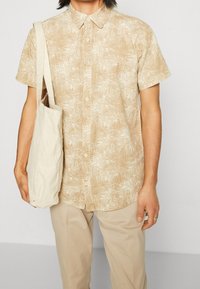 Chemise à col à manches courtes en beige clair avec un motif de feuilles. Sac à main en tissu blanc cassé. Mannequin portant un pantalon kaki, se tenant devant un fond neutre.