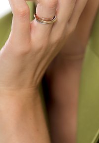 Main avec une bague torsadée tricolore au majeur, reposant près du visage sur un fond de tissu vert.