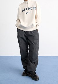 Lichtbeige sweatshirt met halve rits en "NIKE"-logo, gecombineerd met donkergrijze cargobroek met zijzakken. Zwarte stevige laarzen maken de outfit compleet.