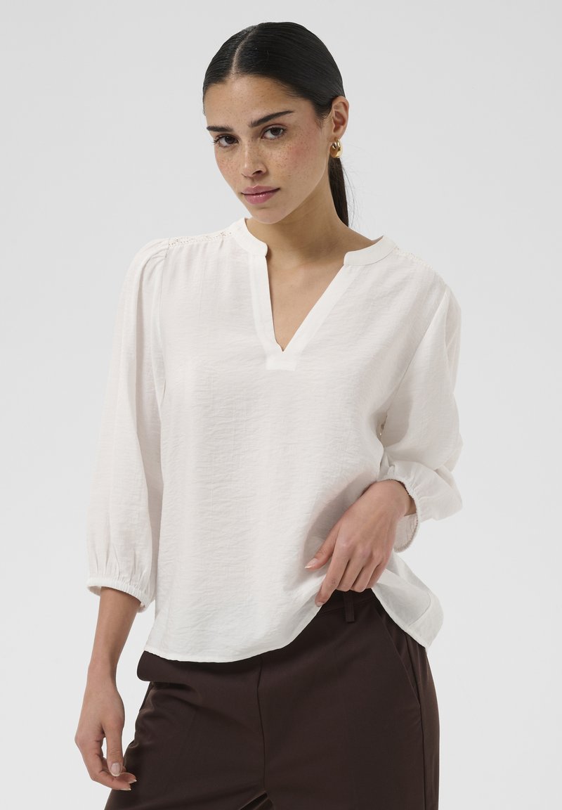Femme aux cheveux foncés portant des boucles d'oreilles créoles dorées, un chemisier blanc à manches longues avec un col en V et un pantalon marron foncé, posant avec la main sur la hanche.
