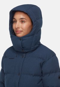 Marinblå isolerad puffjacka med stor huva, försedd med tryckknappar, dragskojusteringar och en logotyp på bröstet.