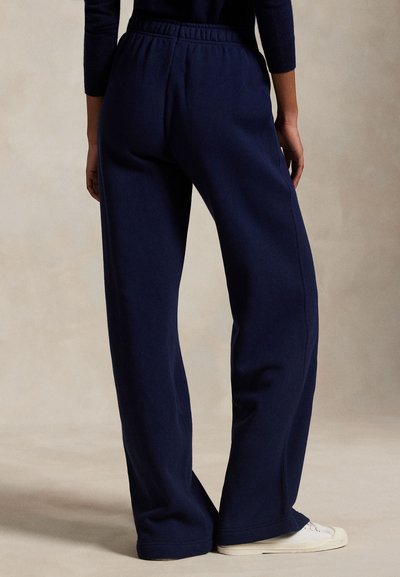 Persoon draagt losse donkerblauwe broek met elastische tailleband en witte sneakers, staat voor een effen beige achtergrond.