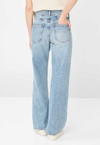 Persona che indossa jeans a zampa d'elefante azzurri chiaro e scarpe bianche, vista da dietro con una mano nella tasca posteriore.