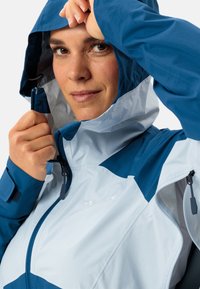 Giacca impermeabile leggera bicolore blu con chiusura lampo frontale, cappuccio regolabile e tasche laterali con lampo, caratterizzata da una texture liscia.