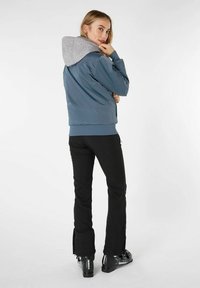 Una giacca blu con zip e cappuccio grigio, dotata di polsini e orlo elasticizzati, abbinata a pantaloni neri e scarponi da sci neri.
